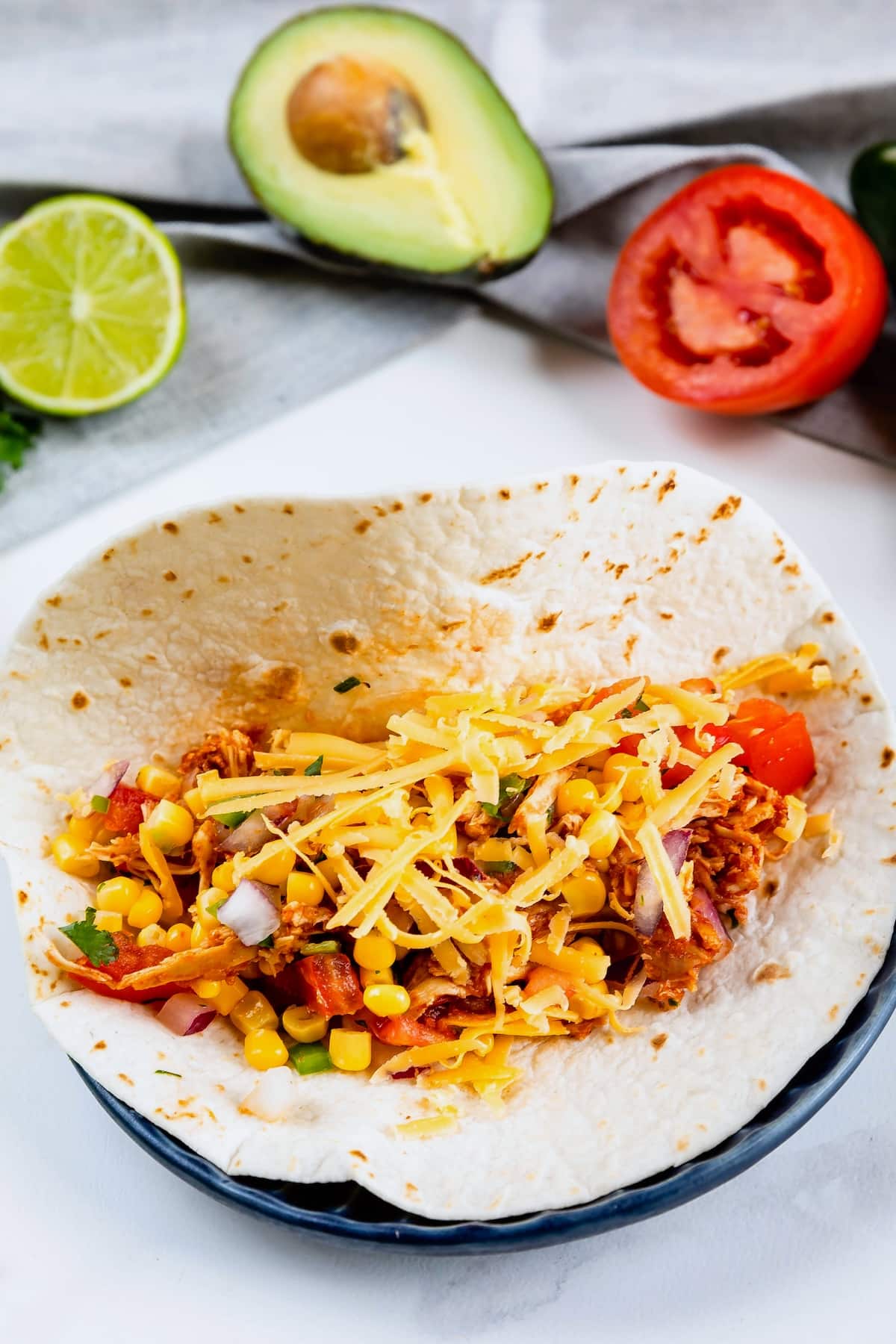 A tortilla filled with shredded chicken, corn, diced tomatoes, onions, and shredded cheese sits on a plate. In the background are half a lime, half an avocado, and a sliced tomato.