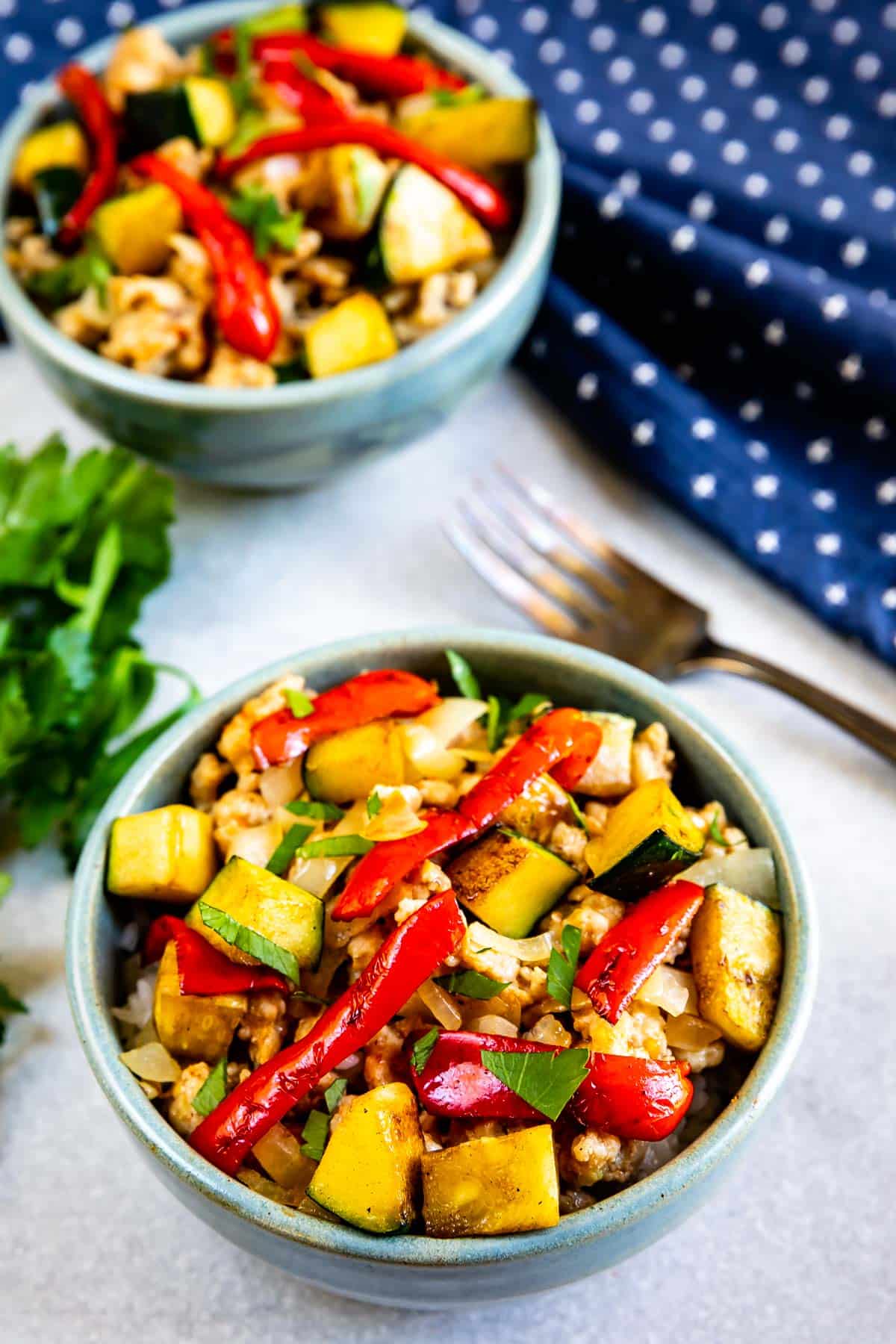 Two bowls filled with a colorful stir-fry featuring zucchini, red bell peppers, chopped greens, and pieces of protein, set on a light surface with a fork, blue polka dot napkin, and fresh herbs nearby.