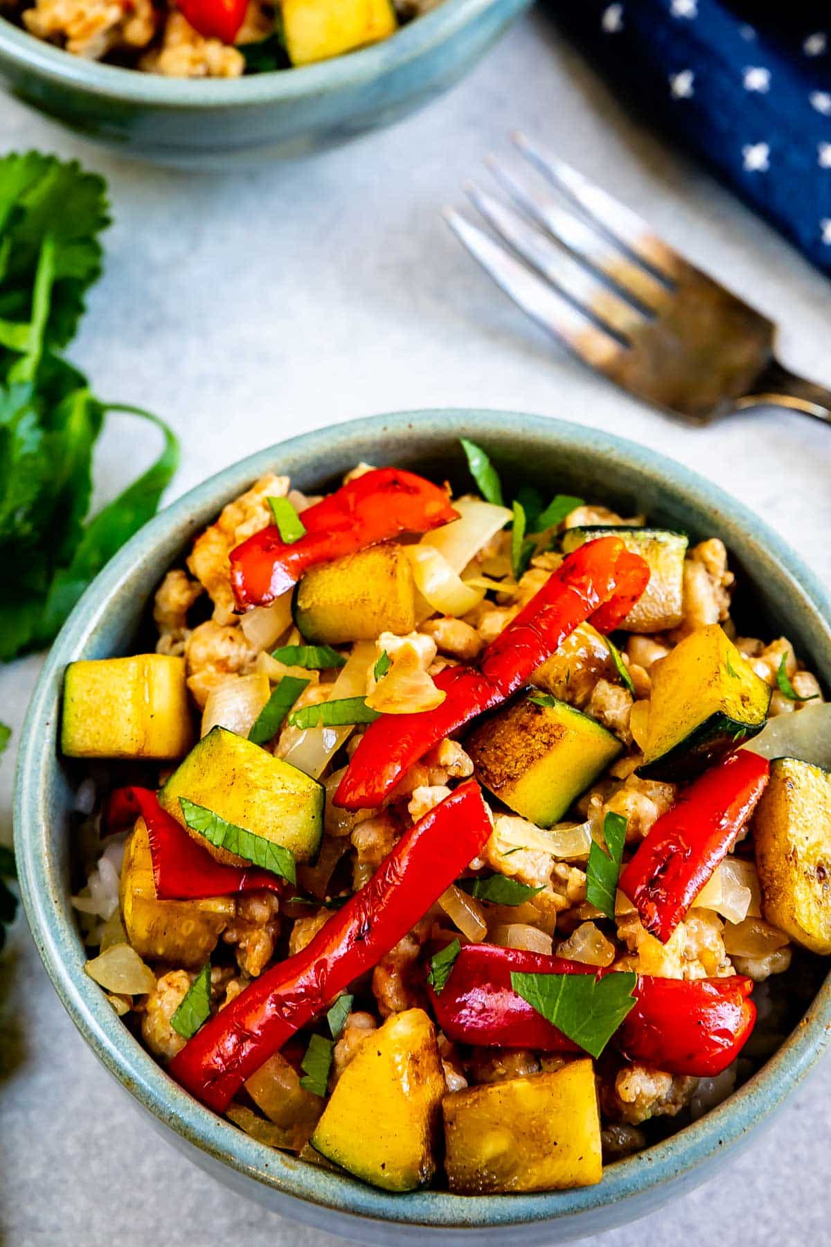 A bowl filled with stir-fried vegetables including zucchini, red bell peppers, and onions, garnished with fresh herbs. A fork and a sprig of cilantro are nearby on a light surface.