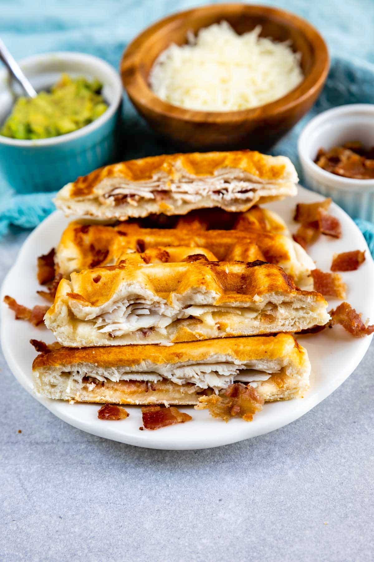 A plate with sliced waffle sandwiches filled with turkey and bacon, with small bowls of shredded cheese, bacon pieces, and guacamole in the background.