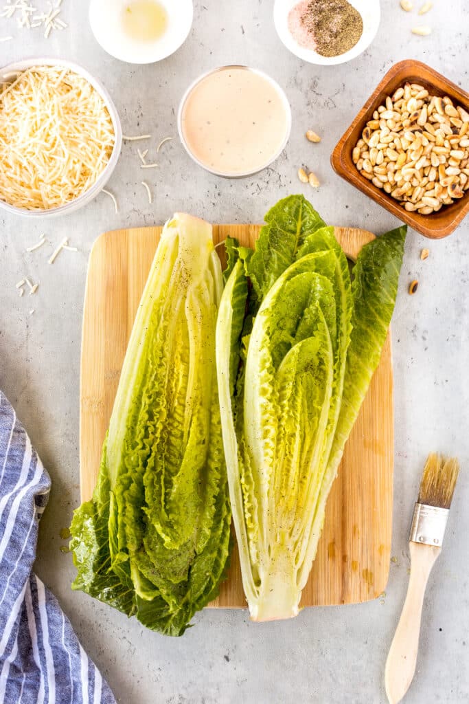 Overhead view of romaine lettuce halves on a wooden board, surrounded by shredded cheese, dressing, pine nuts, spices, a basting brush, and a blue towel on a light countertop.