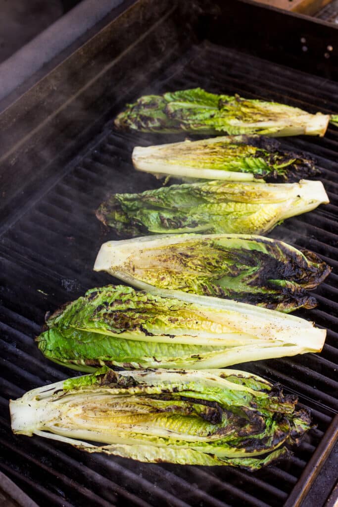 Halved romaine lettuce heads being grilled on a barbecue, with light char marks and steam rising from the greens.