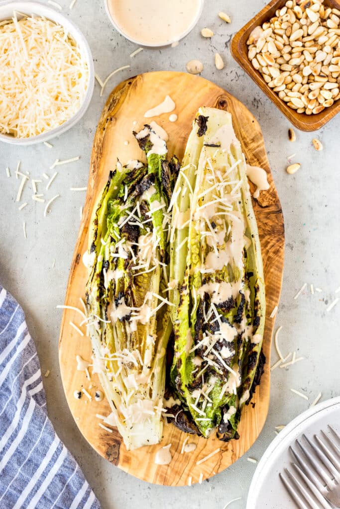 Grilled romaine lettuce halves topped with Caesar dressing and shredded parmesan cheese, served on a wooden board. Surrounding the board are bowls of parmesan, dressing, pine nuts, and a striped napkin.