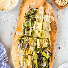 Grilled romaine lettuce topped with shredded cheese, pine nuts, and creamy dressing on a wooden serving board, surrounded by bowls of cheese, pine nuts, a napkin, and a plate with forks.