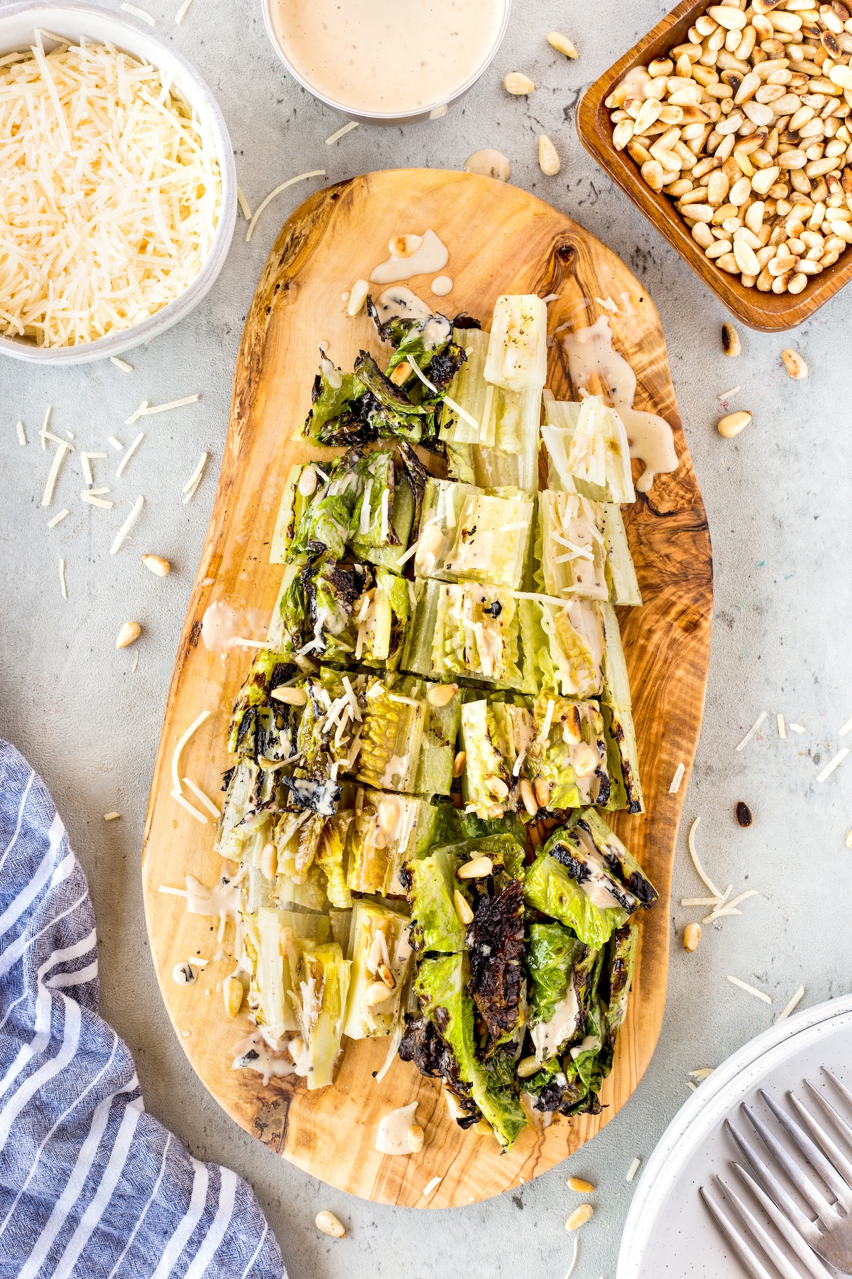 Grilled romaine lettuce topped with shredded cheese, pine nuts, and creamy dressing on a wooden serving board, surrounded by bowls of cheese, pine nuts, a napkin, and a plate with forks.