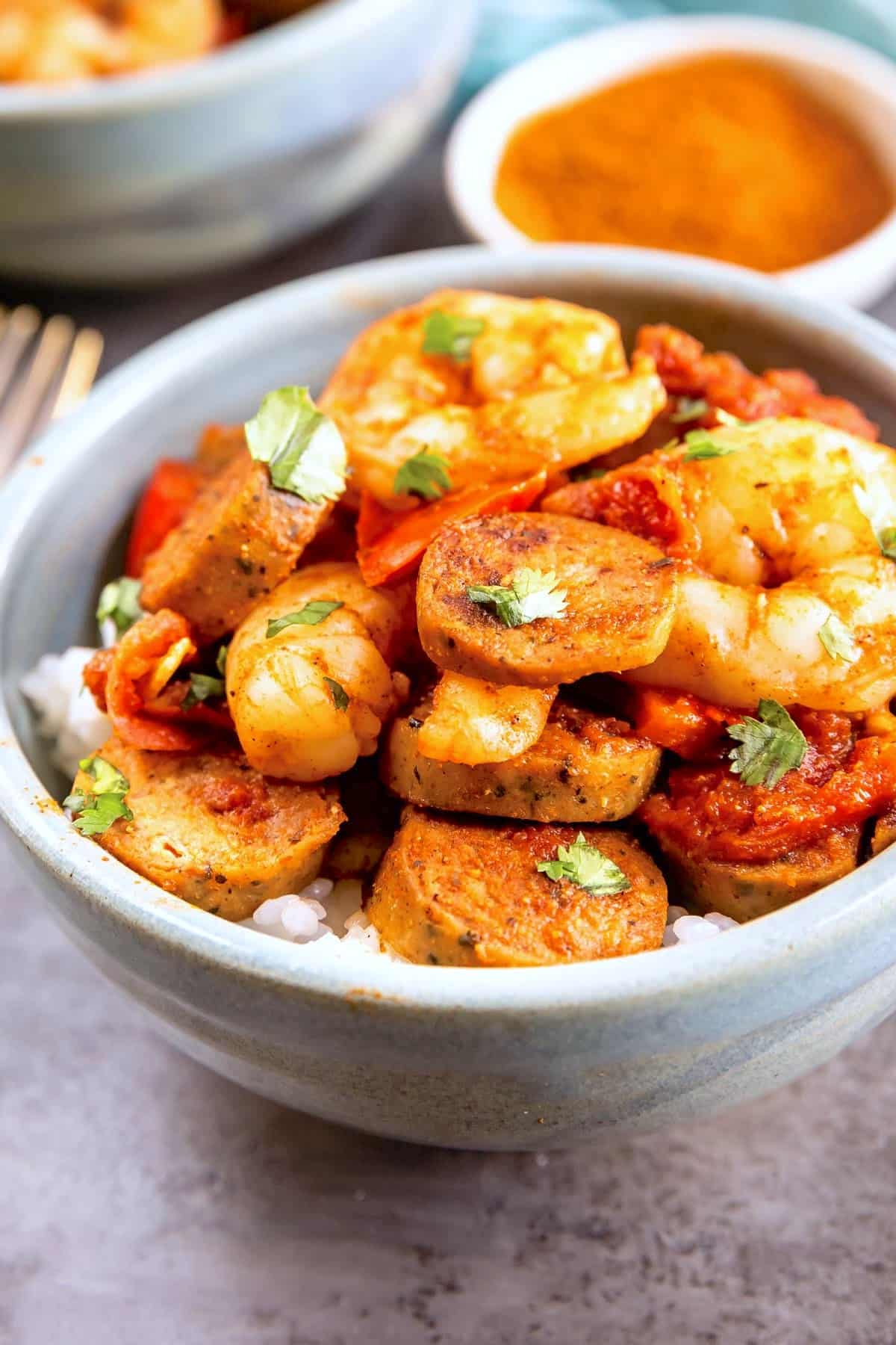 A close-up of a bowl filled with white rice, topped with sliced sausage, shrimp, tomato-based sauce, and garnished with chopped cilantro. A small dish of spices is blurred in the background.