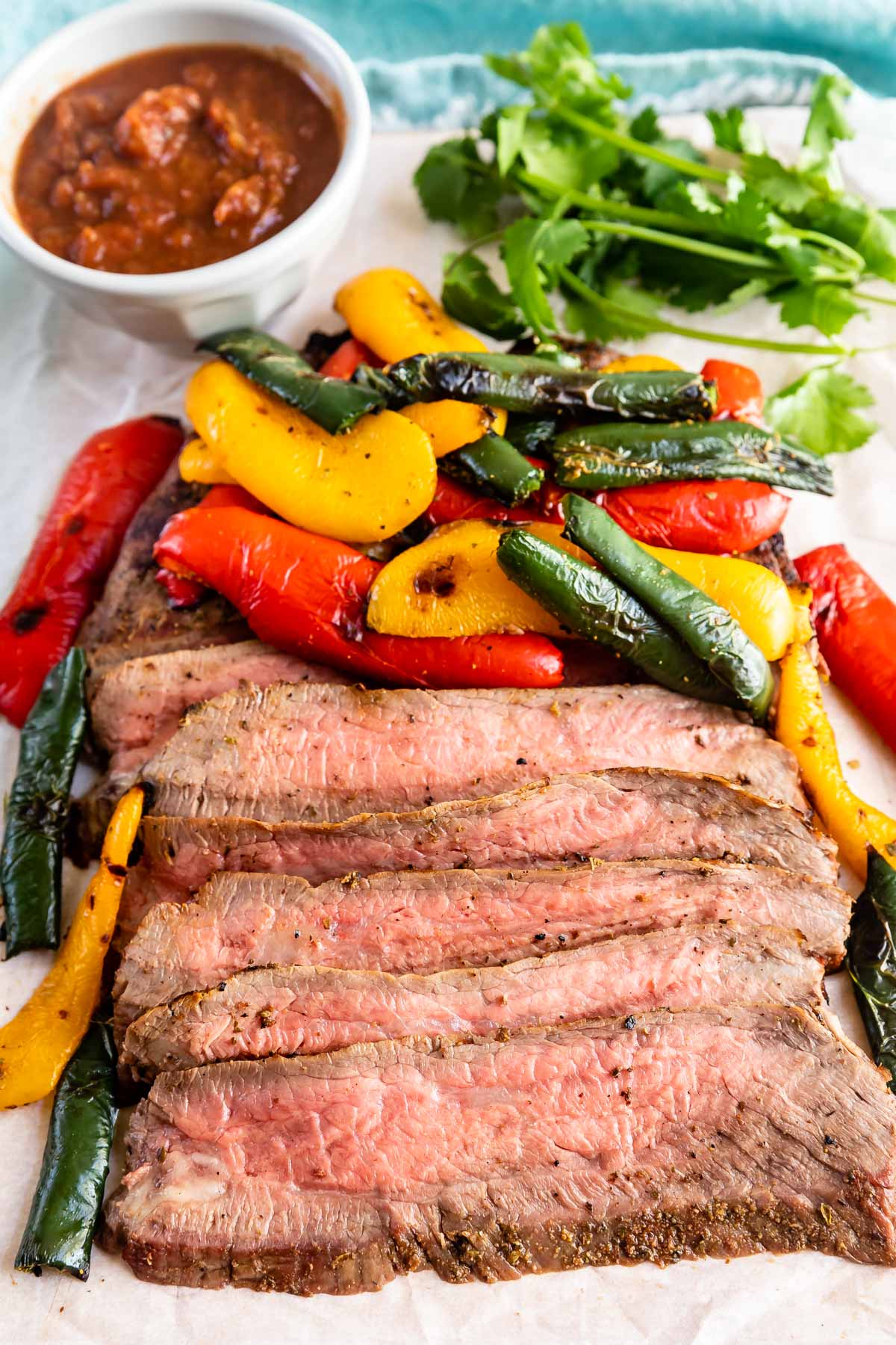 Sliced grilled steak topped with charred red, yellow, and green bell peppers, served on parchment paper with a side of salsa and fresh cilantro in the background.