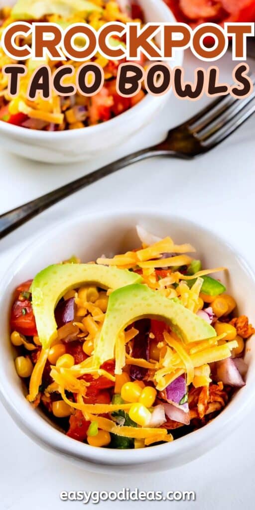 A white bowl filled with a colorful crockpot taco bowl, topped with sliced avocado, shredded cheese, corn, red onion, and tomatoes. A fork and another bowl are visible in the background. Text overlay reads “Crockpot Taco Bowls.”.