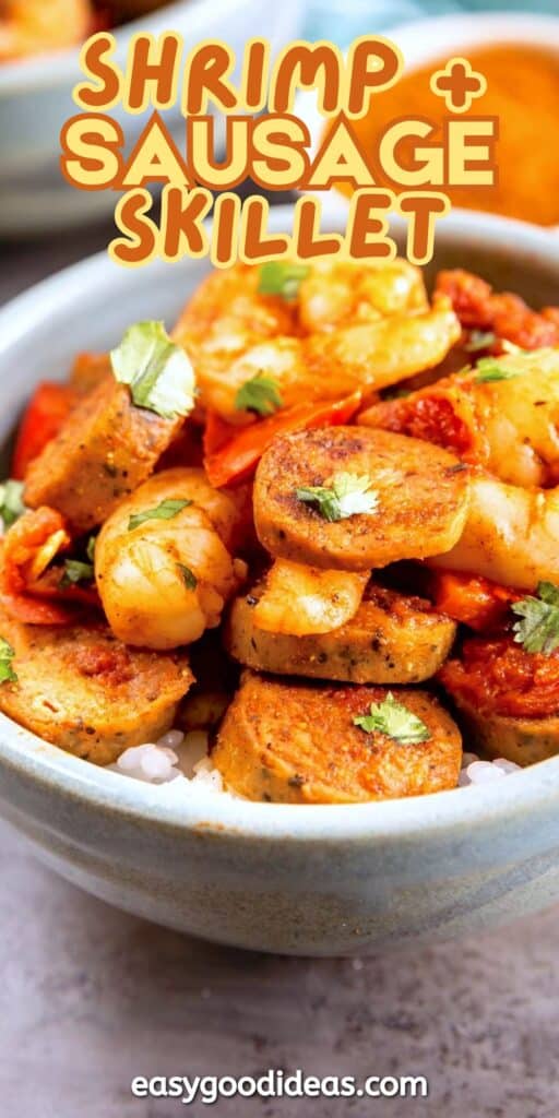 A close-up of a bowl filled with shrimp, sliced sausage, and a tomato-based sauce, garnished with fresh herbs. Text over the image reads Shrimp + Sausage Skillet and easygoodideas.com at the bottom.