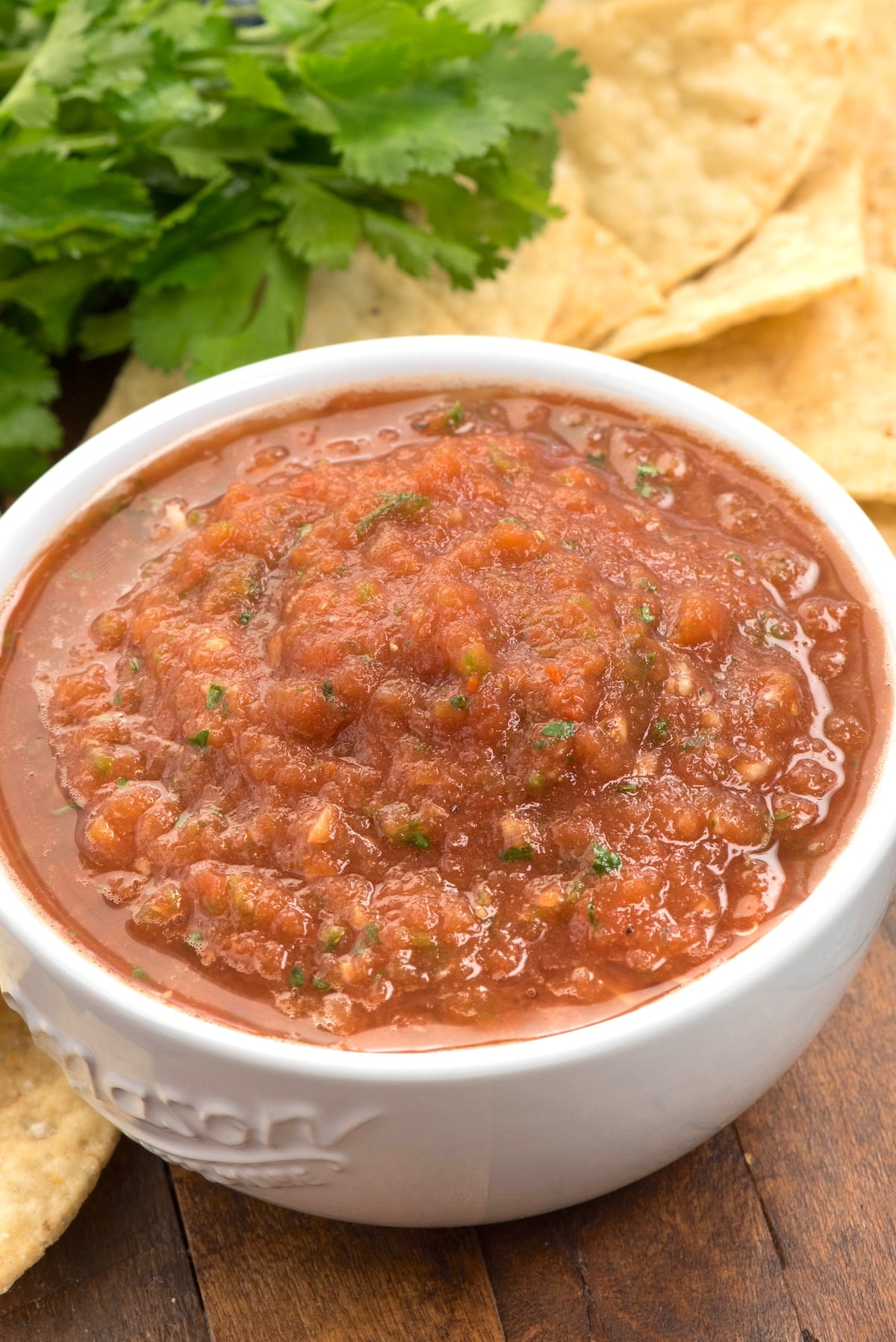 A white bowl filled with chunky red salsa sits on a wooden surface, surrounded by tortilla chips and fresh cilantro in the background.