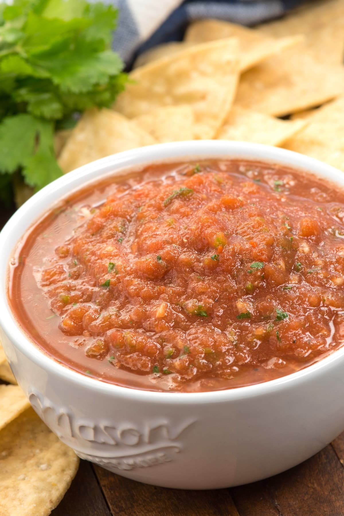 A white bowl filled with chunky red salsa sits on a wooden surface, surrounded by tortilla chips and fresh cilantro in the background.