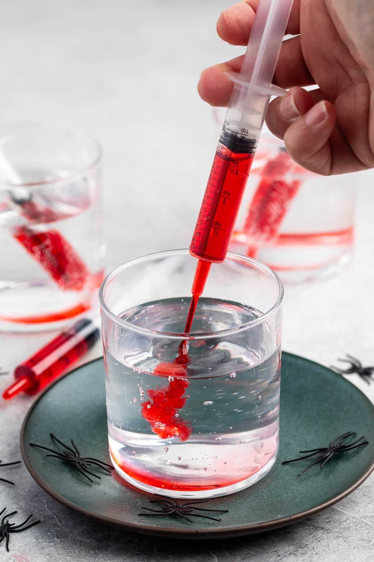 A hand holds a syringe filled with red liquid, injecting it into a clear glass of water on a green plate decorated with small plastic black spiders. Another glass and syringe are in the background.