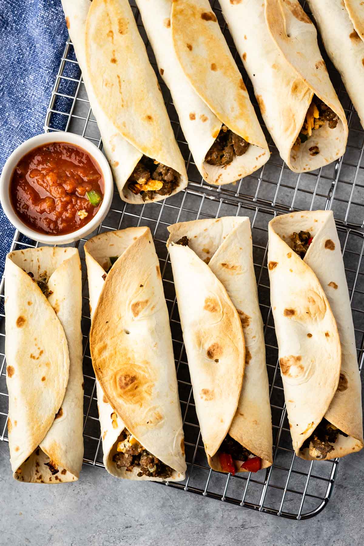 Baked tortillas filled with meat and vegetables are arranged on a wire rack. A small bowl of salsa is placed on the side, and a blue cloth is partially visible in the corner.