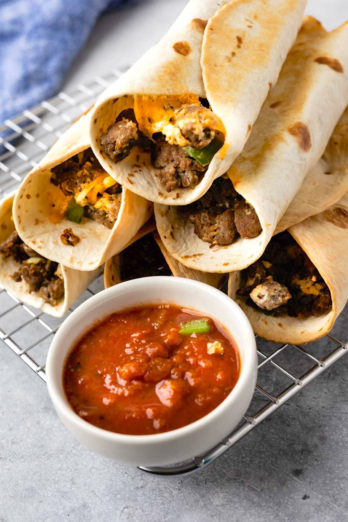 A stack of beef and cheese taquitos on a wire rack, with a small bowl of chunky red salsa in front. The taquitos are wrapped in golden-brown tortillas and filled with ground beef and melted cheese.