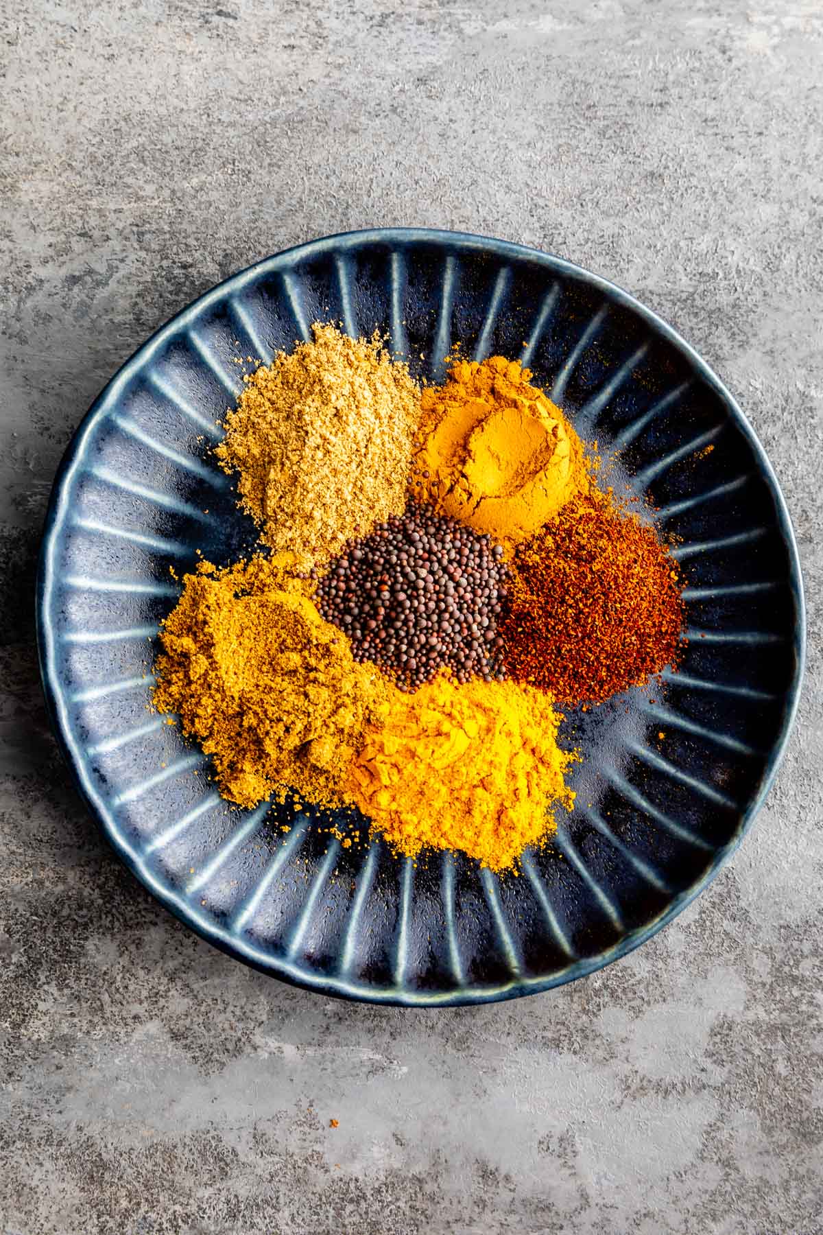 A dark blue plate with five different colorful ground spices and whole mustard seeds arranged in small piles on a gray textured surface.