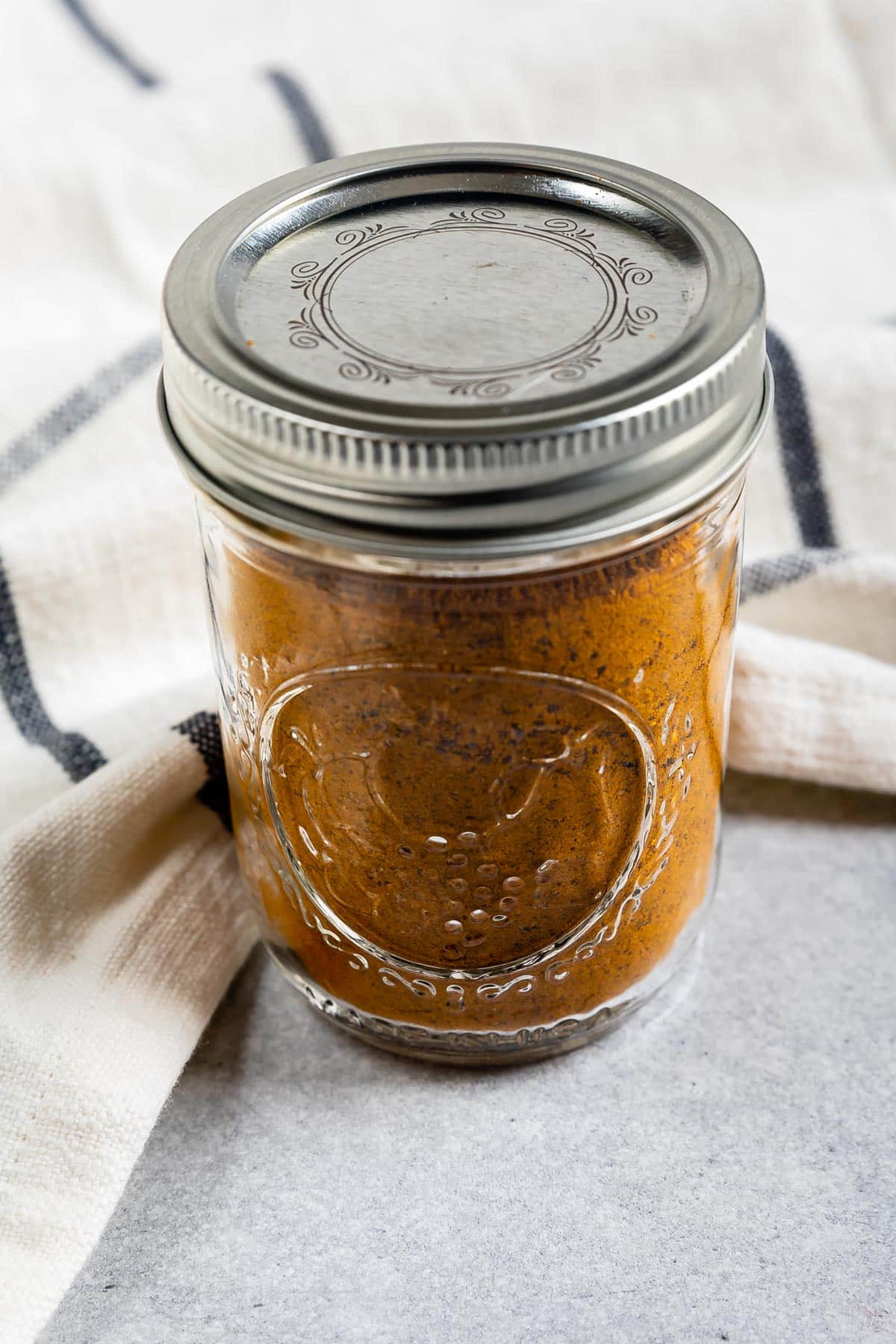 A glass mason jar filled with a reddish-brown spice blend, sealed with a metal lid, sits on a light gray surface next to a white and blue striped kitchen towel.
