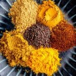 A dark plate holding six colorful piles of spices, including yellow turmeric, brown mustard seeds, ground chili, curry powder, coriander, and cumin, arranged in a circle.