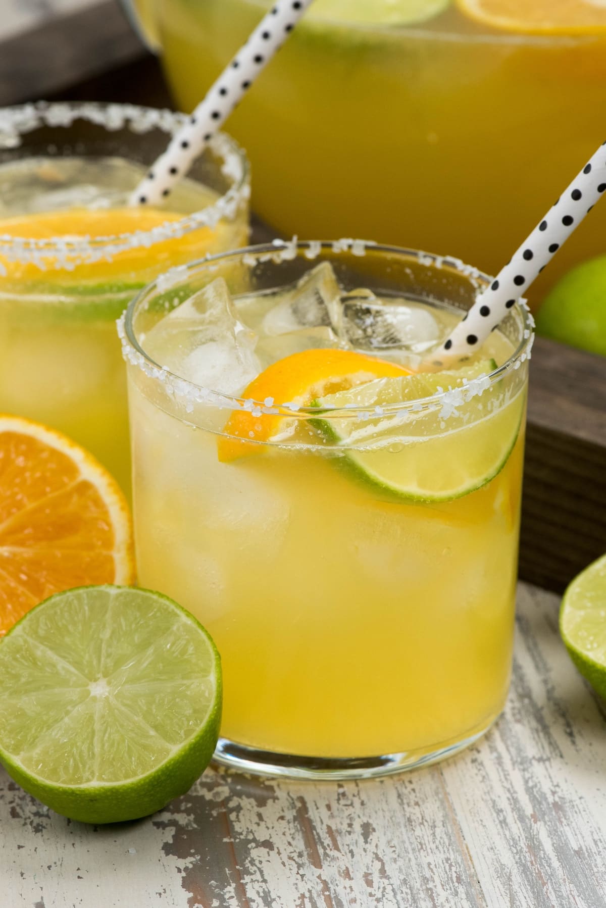 Two glasses of a citrus cocktail with ice, garnished with lime and orange slices, and salt-rimmed edges. Black and white polka dot straws are in each glass, with whole and sliced citrus fruits nearby.