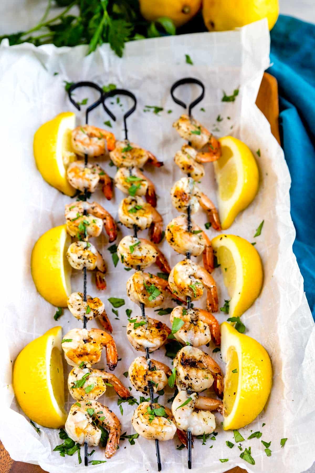 Grilled shrimp skewers garnished with chopped herbs are arranged on parchment paper, surrounded by large lemon wedges and fresh parsley in the background.