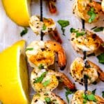 Grilled shrimp skewers garnished with chopped herbs are arranged on parchment paper next to two lemon wedges.