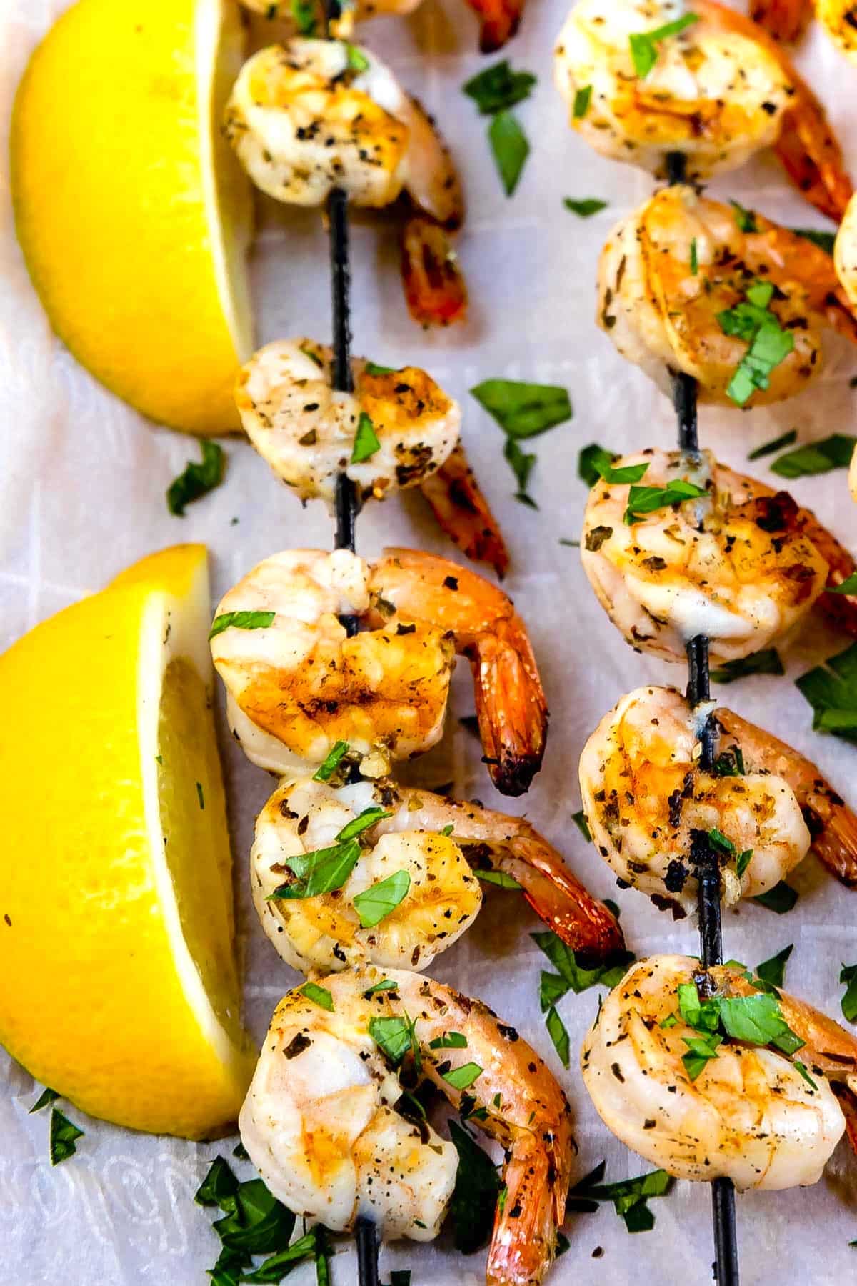 Grilled shrimp skewers garnished with chopped herbs are arranged on parchment paper next to two lemon wedges.