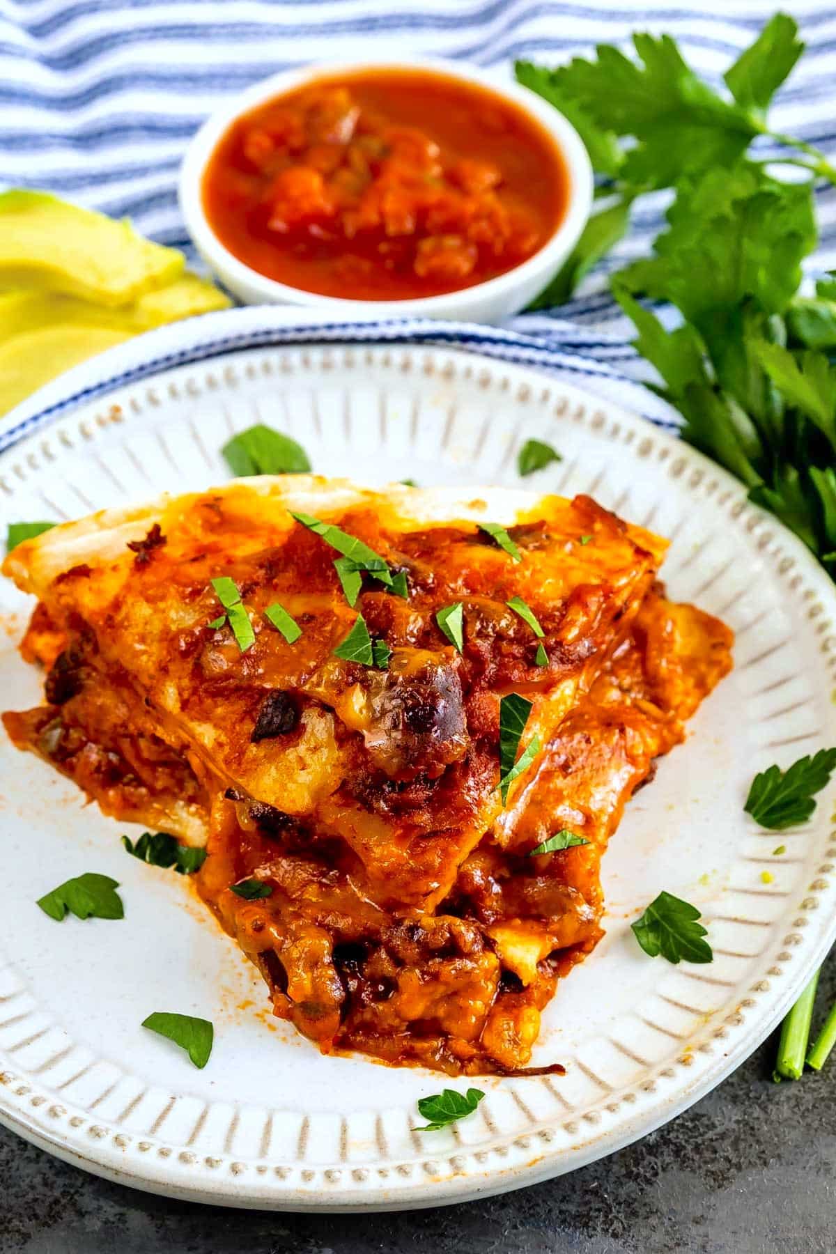 A serving of cheesy, baked lasagna topped with fresh parsley sits on a white plate. A bowl of tomato sauce and fresh parsley are in the background.