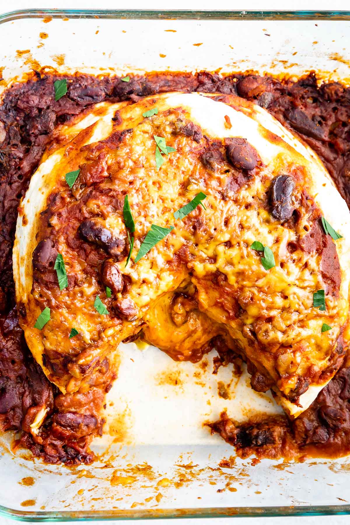A baked tortilla pie filled with beans, cheese, and sauce, topped with melted cheese and garnished with chopped herbs, sits in a glass baking dish with a wedge missing.