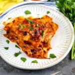 A slice of cheesy enchilada casserole garnished with fresh herbs on a white plate, next to a fork, with a bowl of salsa and fresh cilantro in the background.