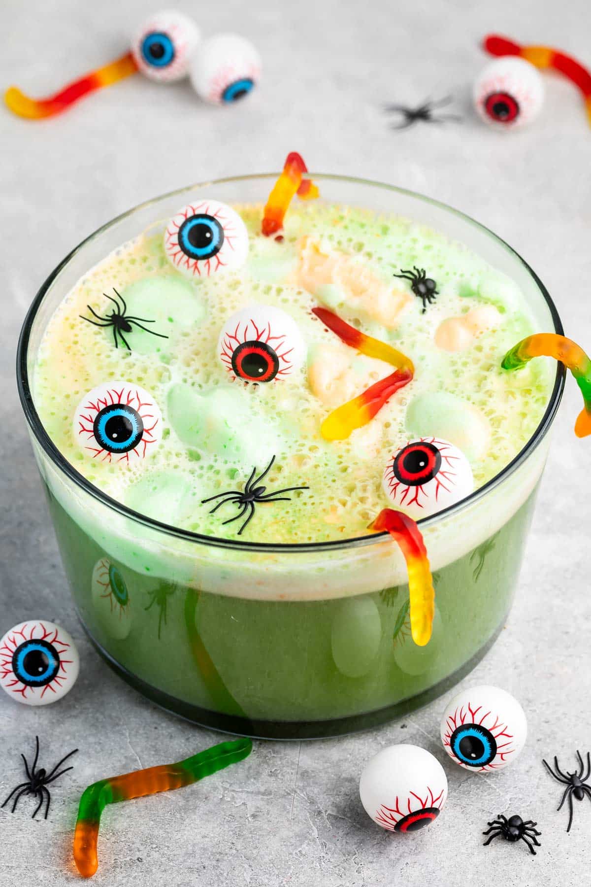A green punch bowl filled with floating marshmallows, gummy worms, plastic eyeballs, and fake spiders, creating a spooky Halloween-themed drink. More eyeballs and spiders are scattered on the tabletop around the bowl.