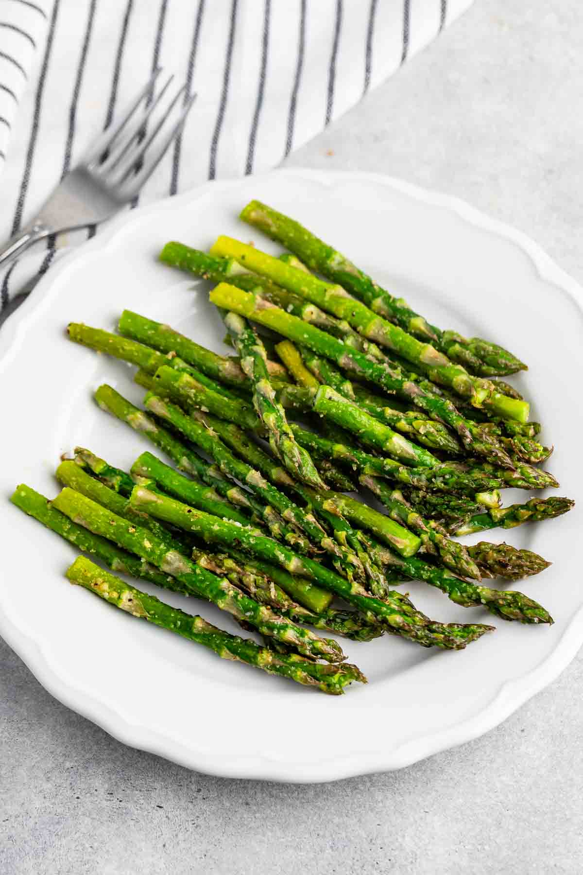 A white plate filled with cooked asparagus spears seasoned with spices and herbs, set on a light gray surface next to a striped cloth napkin and a fork.