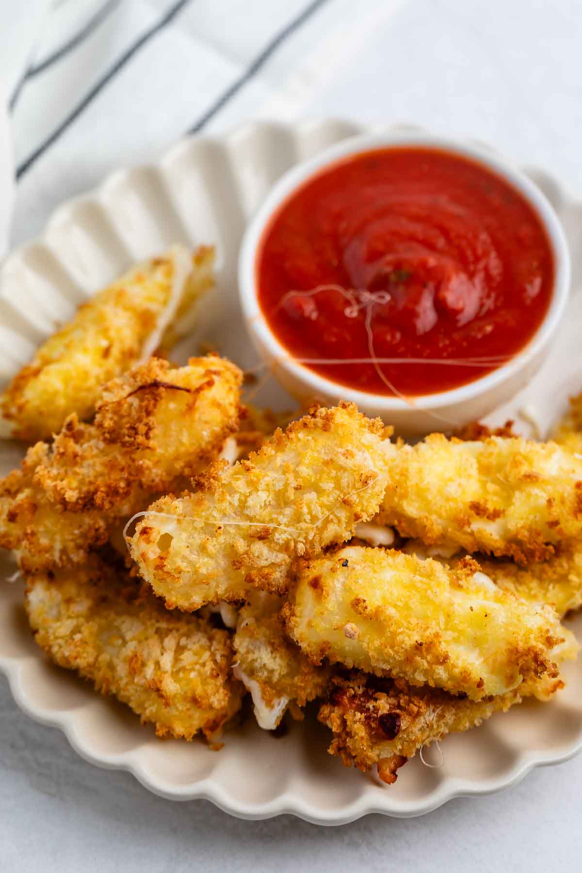 A plate of crispy, golden-brown mozzarella sticks is served with a small bowl of marinara sauce for dipping. The mozzarella sticks are arranged neatly on a cream-colored, scalloped plate.