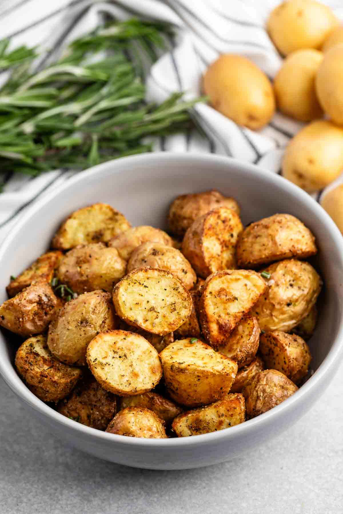 A gray bowl filled with golden roasted potato halves, seasoned with herbs, sits on a light surface. Fresh rosemary and whole potatoes are in the background.
