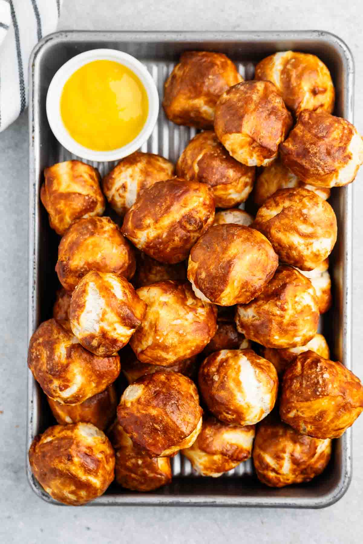 A tray filled with golden-brown pretzel bites, served with a small bowl of yellow dipping sauce on the side. The pretzel bites have a shiny, crisp exterior and are arranged in a metal baking pan.