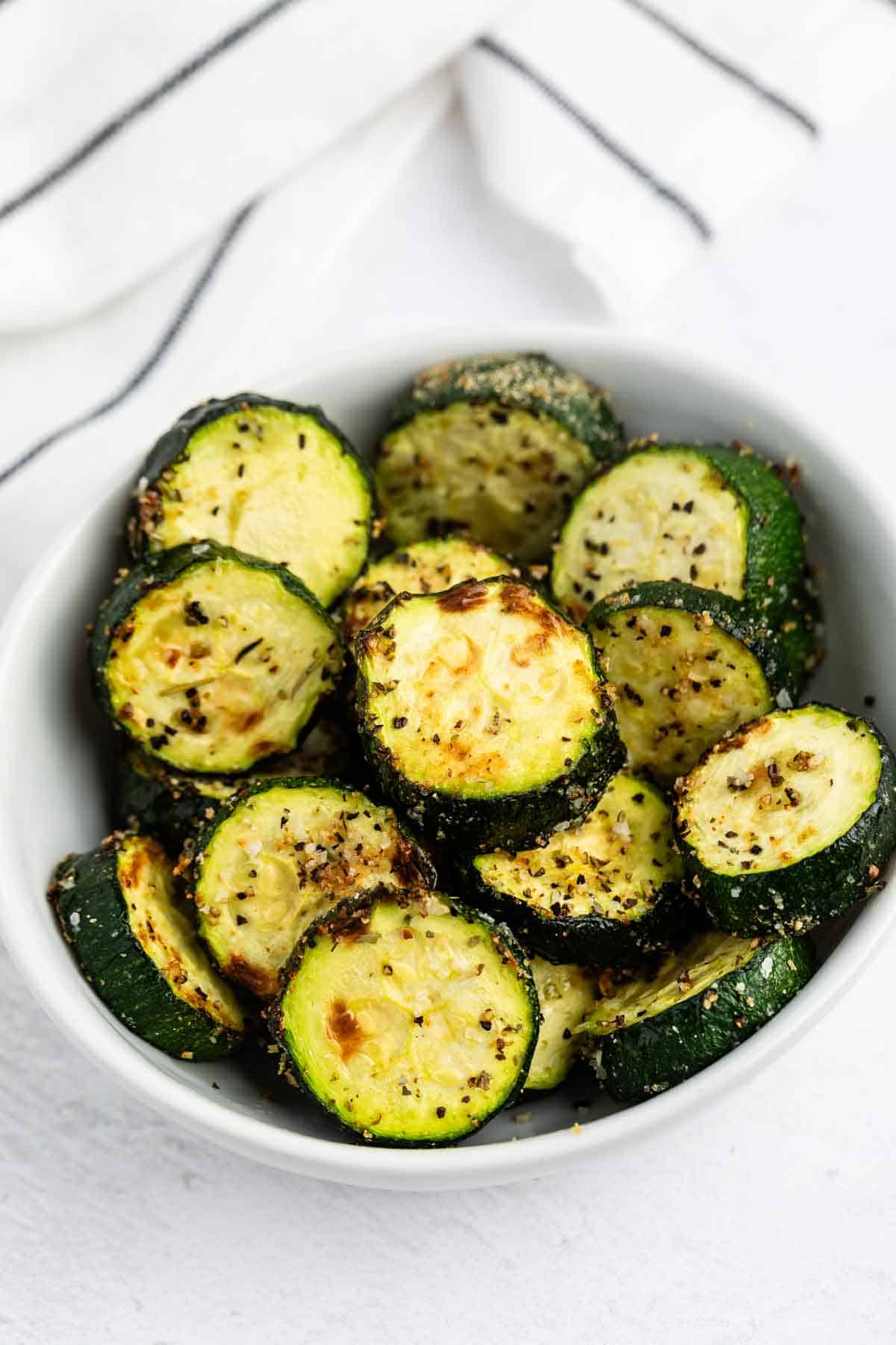 A white bowl filled with roasted zucchini slices seasoned with herbs and spices, sitting on a white surface with a striped cloth in the background.