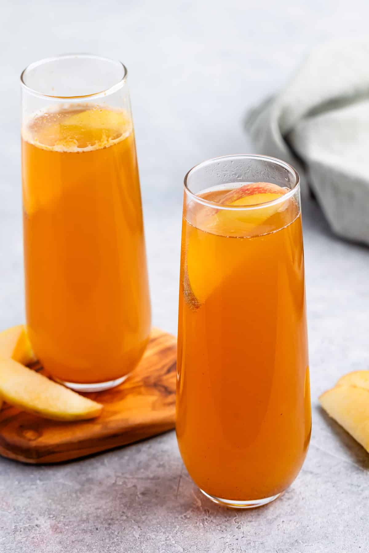 Two tall glasses filled with orange-colored peach iced tea, each garnished with a peach slice. One glass is on a wooden coaster next to peach slices on a light gray surface.