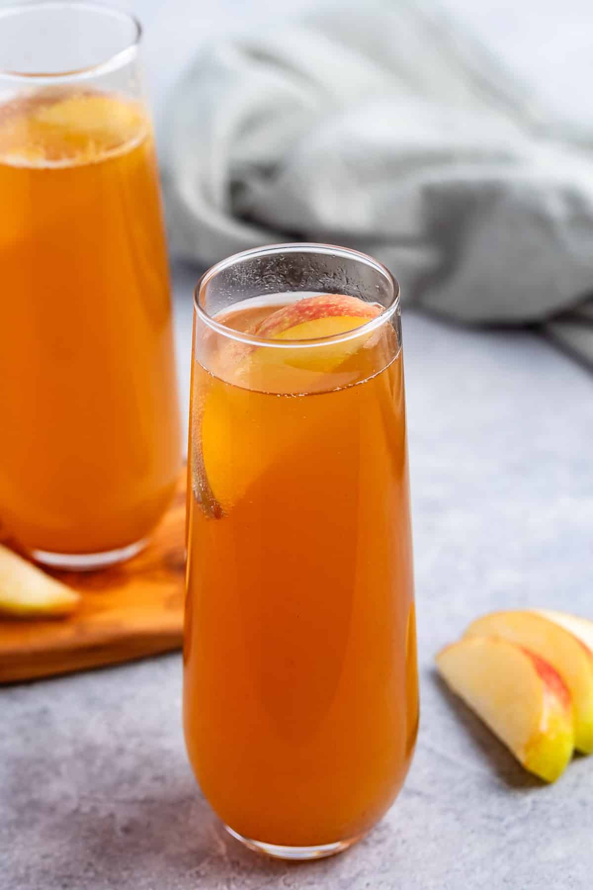 A tall glass of amber-colored apple cider garnished with a fresh apple slice, set on a gray surface with another drink and apple slices in the background.