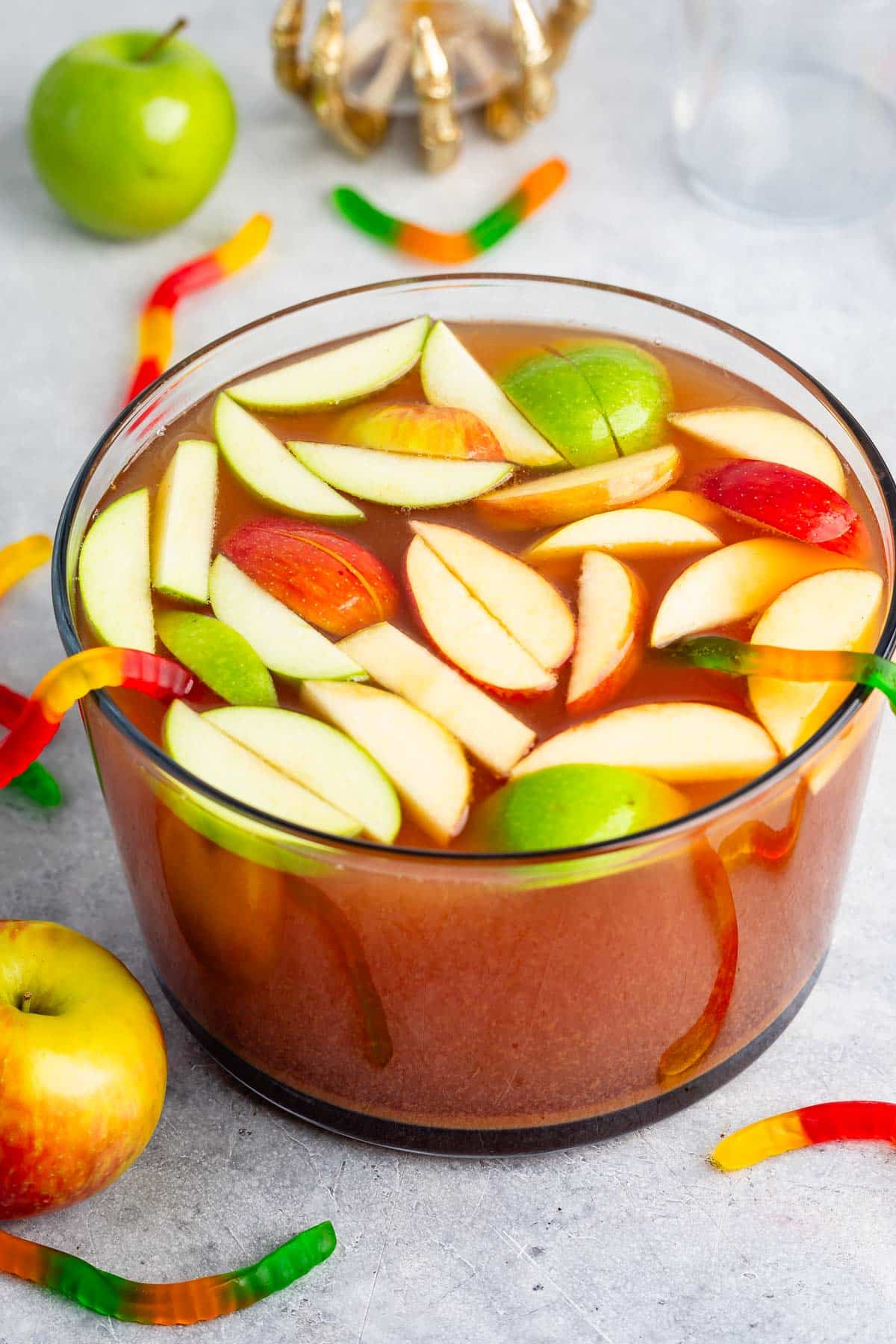 A large glass bowl of punch filled with apple slices and green apples, with colorful gummy worms draped over the edge. Whole apples and gummy worms are scattered on the light gray surface nearby.