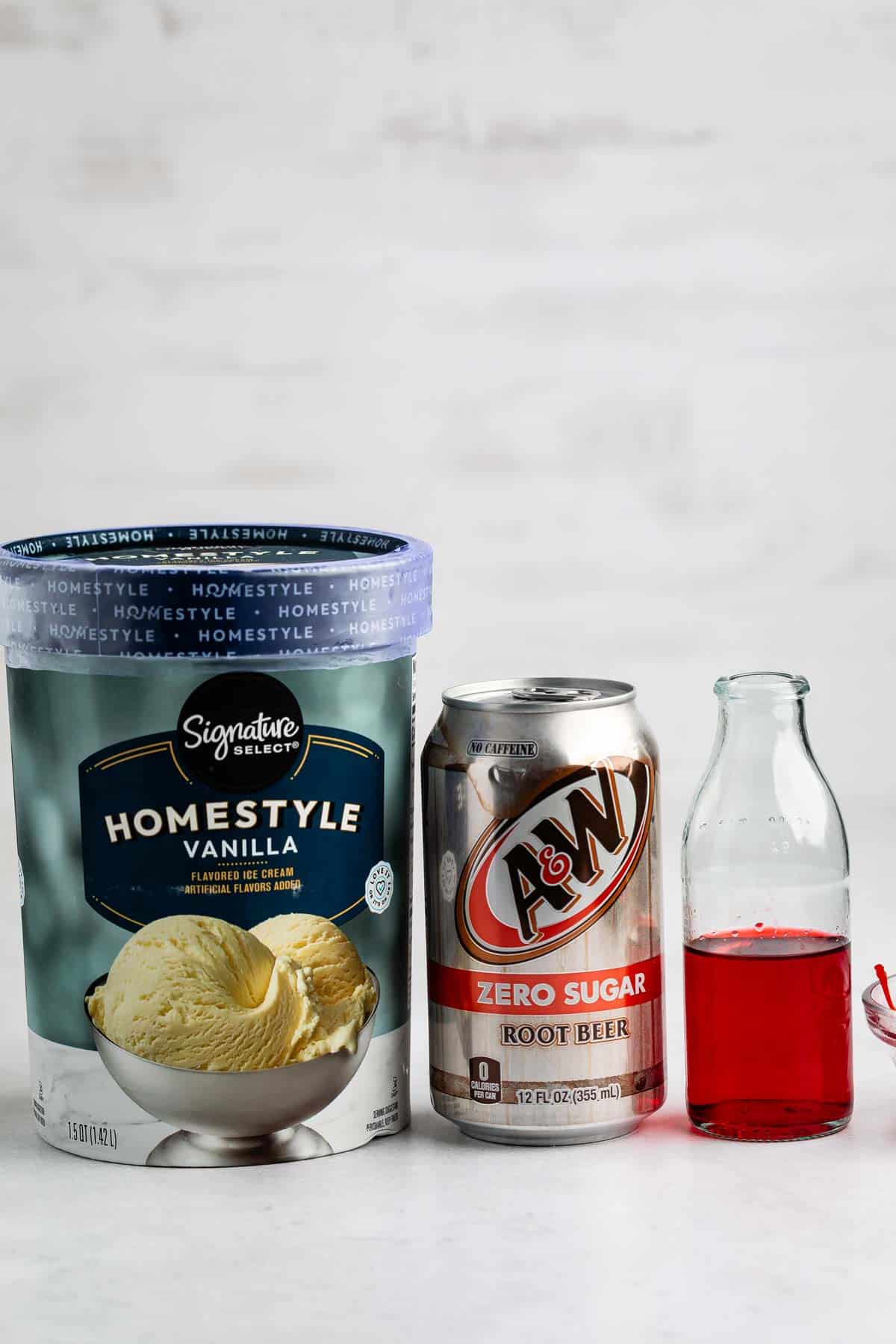 A tub of Signature Select Homestyle Vanilla ice cream, a can of A&W Zero Sugar Root Beer, and a small clear glass bottle filled with red liquid are arranged side by side on a white surface.