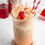 A glass of root beer float with vanilla ice cream, topped with a maraschino cherry and two red-and-white striped straws. An A&W root beer can and a jar of cherries are in the background.