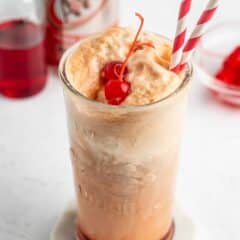 A glass of root beer float with vanilla ice cream, topped with a maraschino cherry and two red-and-white striped straws. An A&W root beer can and a jar of cherries are in the background.