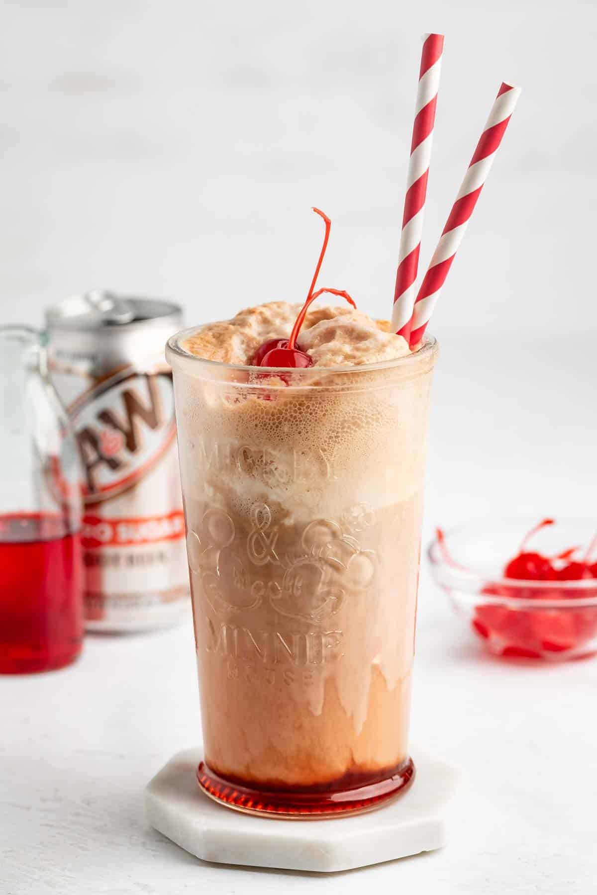 A root beer float with two red-and-white striped straws, topped with foam and a maraschino cherry, sits in a clear glass. A can of root beer and a bowl of cherries are in the background.