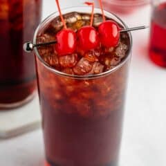A glass of dark soda filled with ice, garnished with three bright red maraschino cherries on a cocktail pick, sits on a white surface. A second glass and a bowl of cherries are blurred in the background.