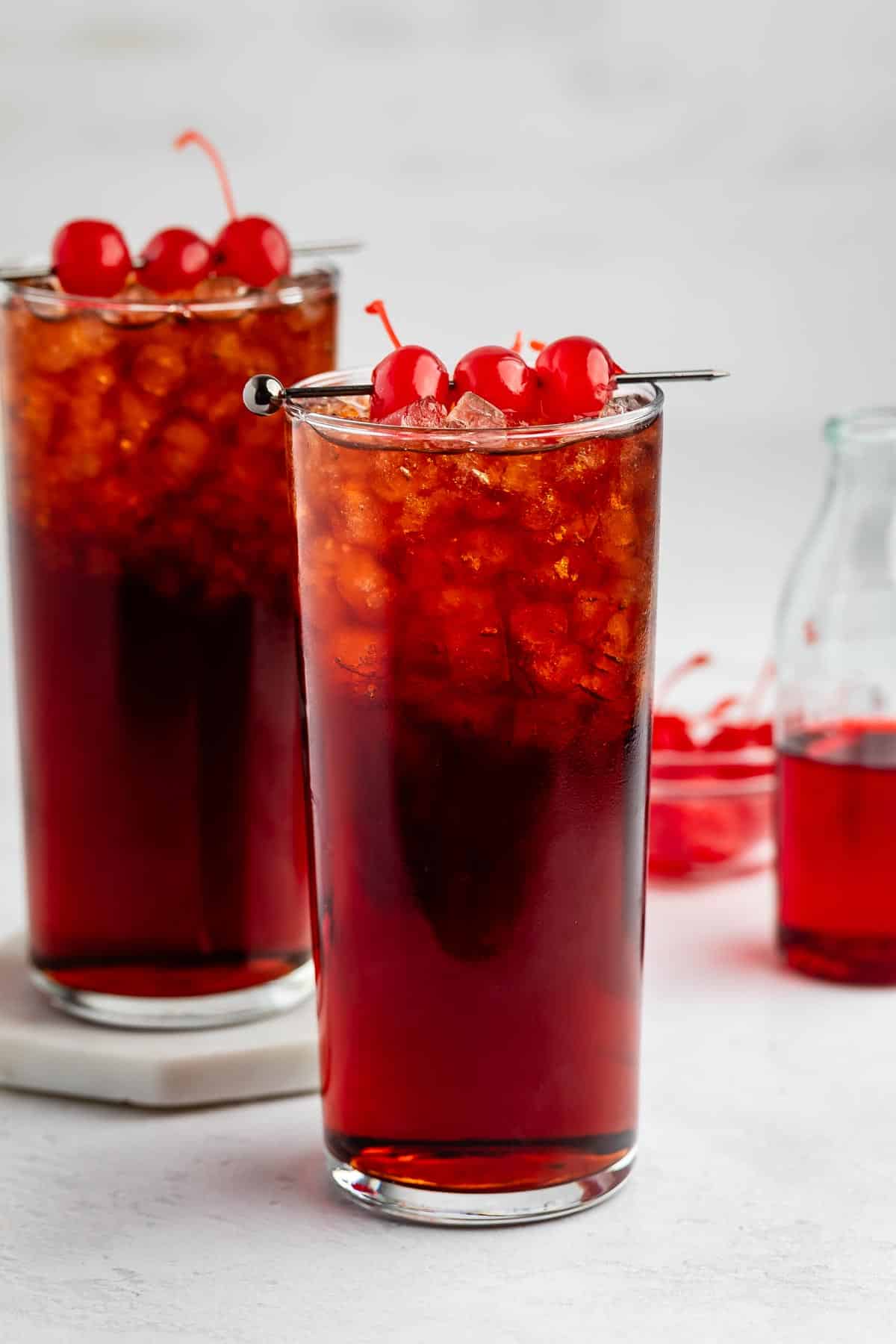 Two tall glasses filled with a dark red iced drink, topped with maraschino cherries and crushed ice. A bowl of cherries and a small bottle of red liquid are in the background on a white surface.