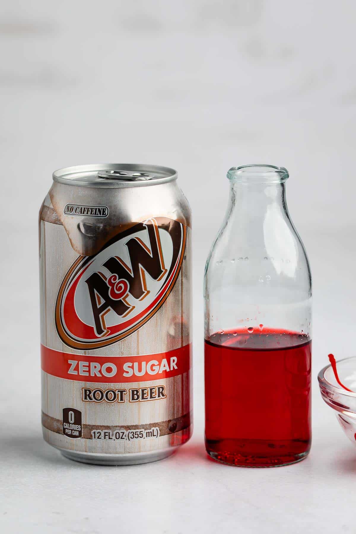 A can of A&W Zero Sugar Root Beer stands next to a small glass bottle containing a red liquid. Part of a glass bowl with red liquid is visible on the right, all set on a white surface.