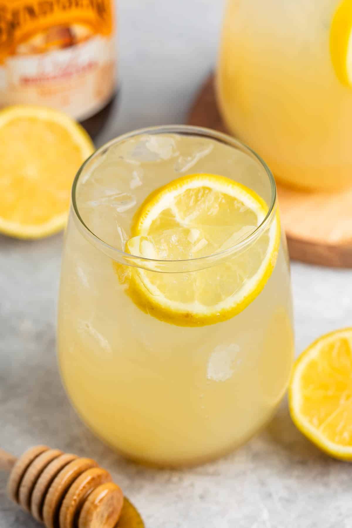 A glass of iced lemonade garnished with a lemon slice sits on a counter. Nearby are lemon halves, a honey dipper with honey, and a pitcher of lemonade in the background.
