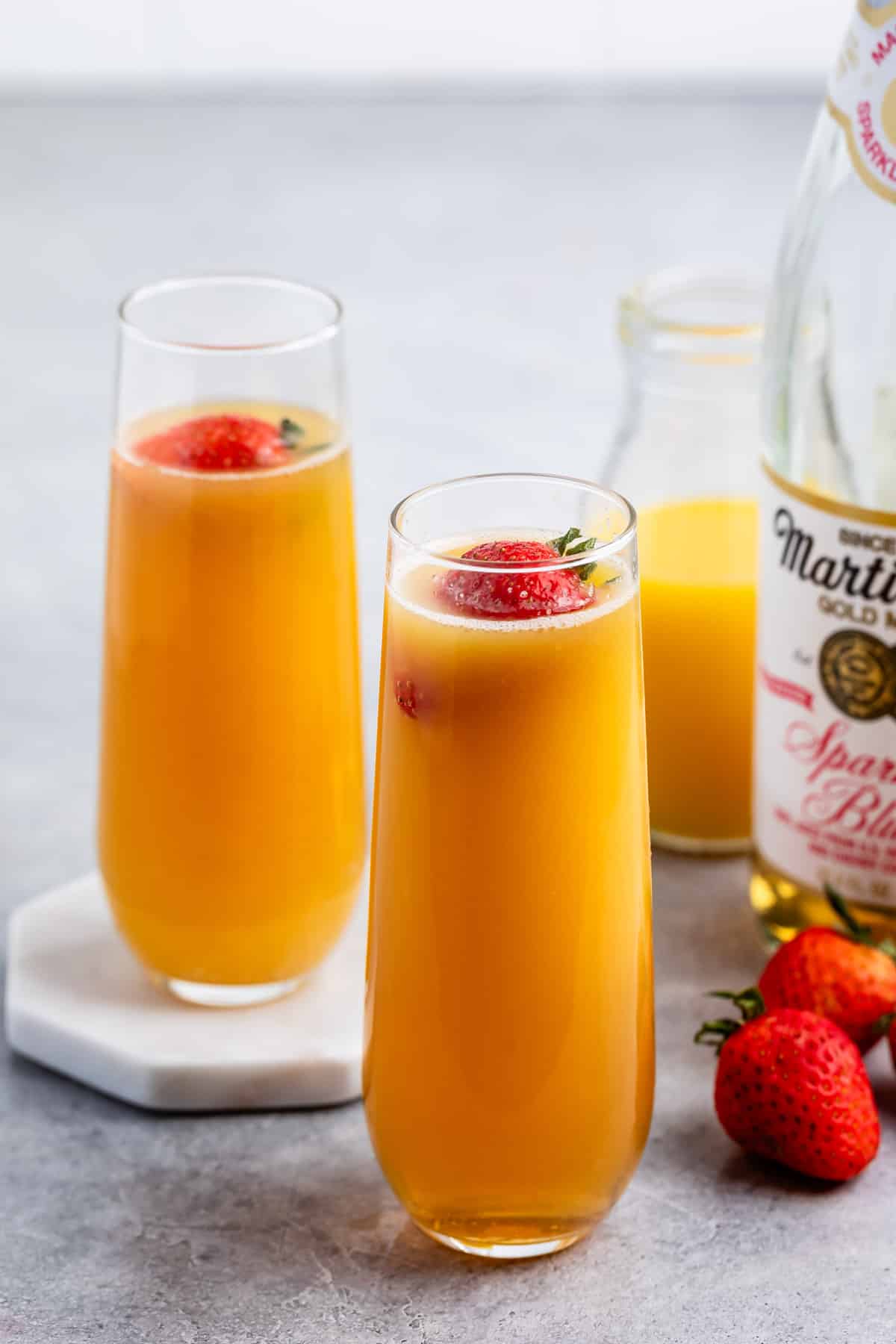 Two tall glasses filled with orange-yellow sparkling juice, each topped with a whole strawberry. A bottle of sparkling juice, a small glass jug with juice, and a few strawberries are in the background.