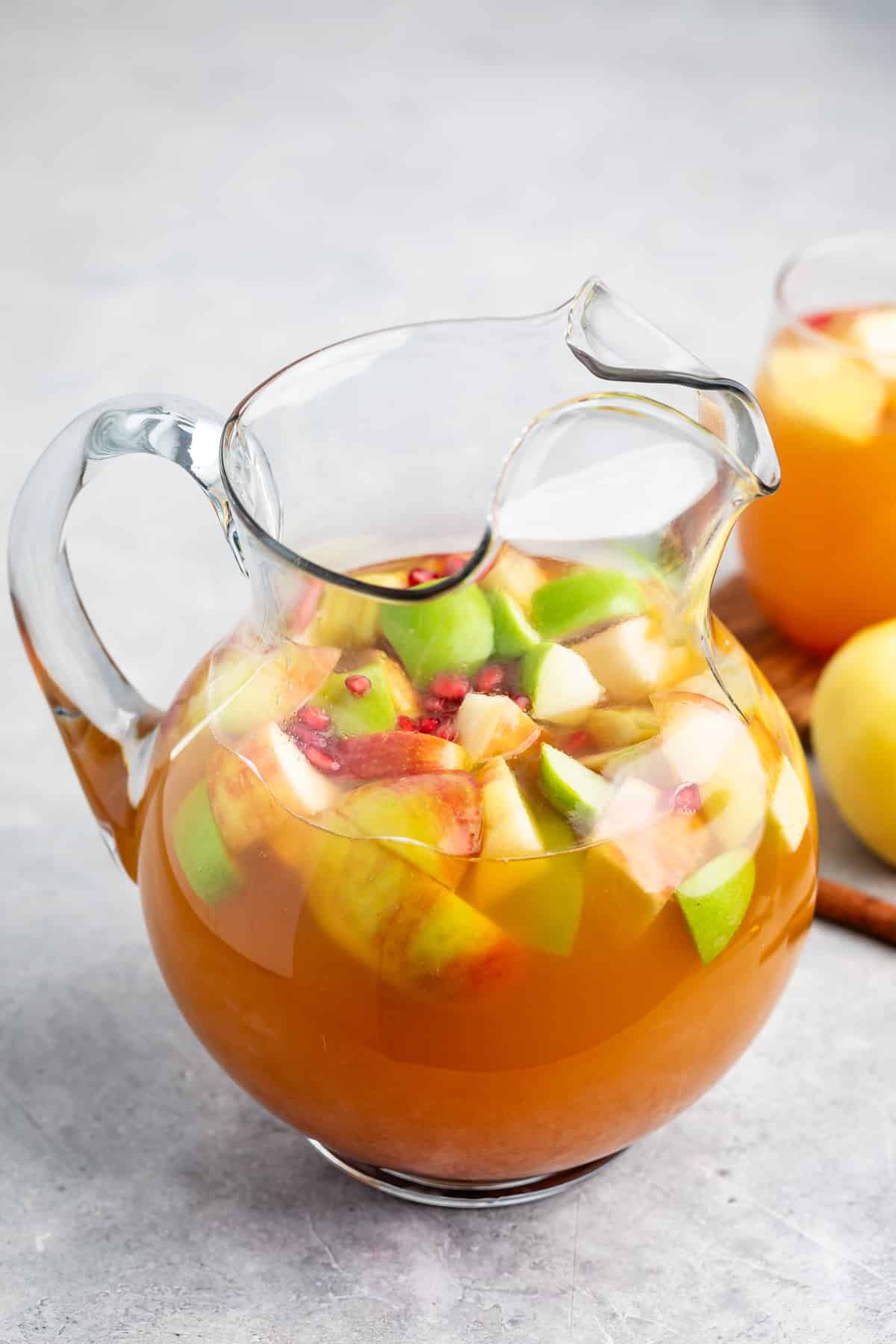 A clear glass pitcher filled with orange-colored punch, containing sliced apples, green apples, and pomegranate seeds, sitting on a light gray surface.