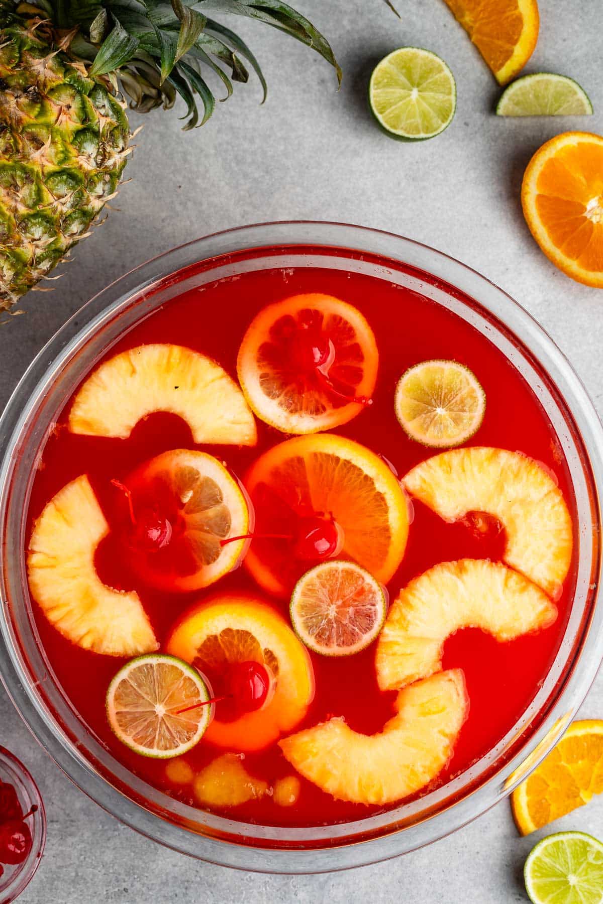 A glass bowl filled with bright red punch, garnished with pineapple rings, orange slices, lime slices, and maraschino cherries, with fresh fruit slices and a pineapple nearby on a gray surface.