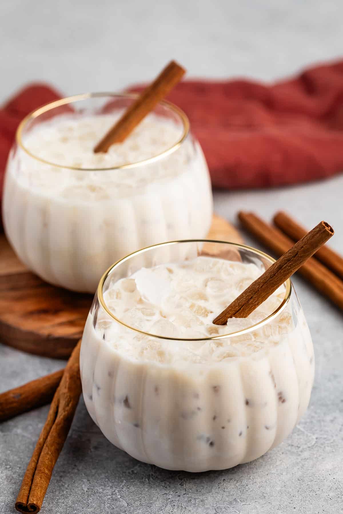 Two round glasses filled with an iced creamy beverage, each garnished with a cinnamon stick. Extra cinnamon sticks and a red cloth are nearby on a light-colored surface.