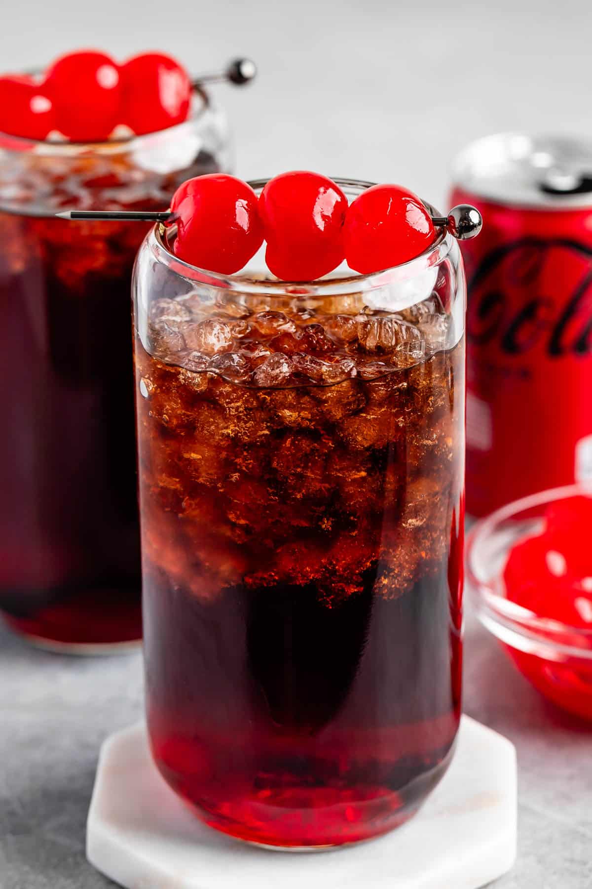 A glass of cherry cola with ice, garnished with three maraschino cherries on top, sits on a coaster. Another similar drink, a bowl of cherries, and a red soda can are in the background.